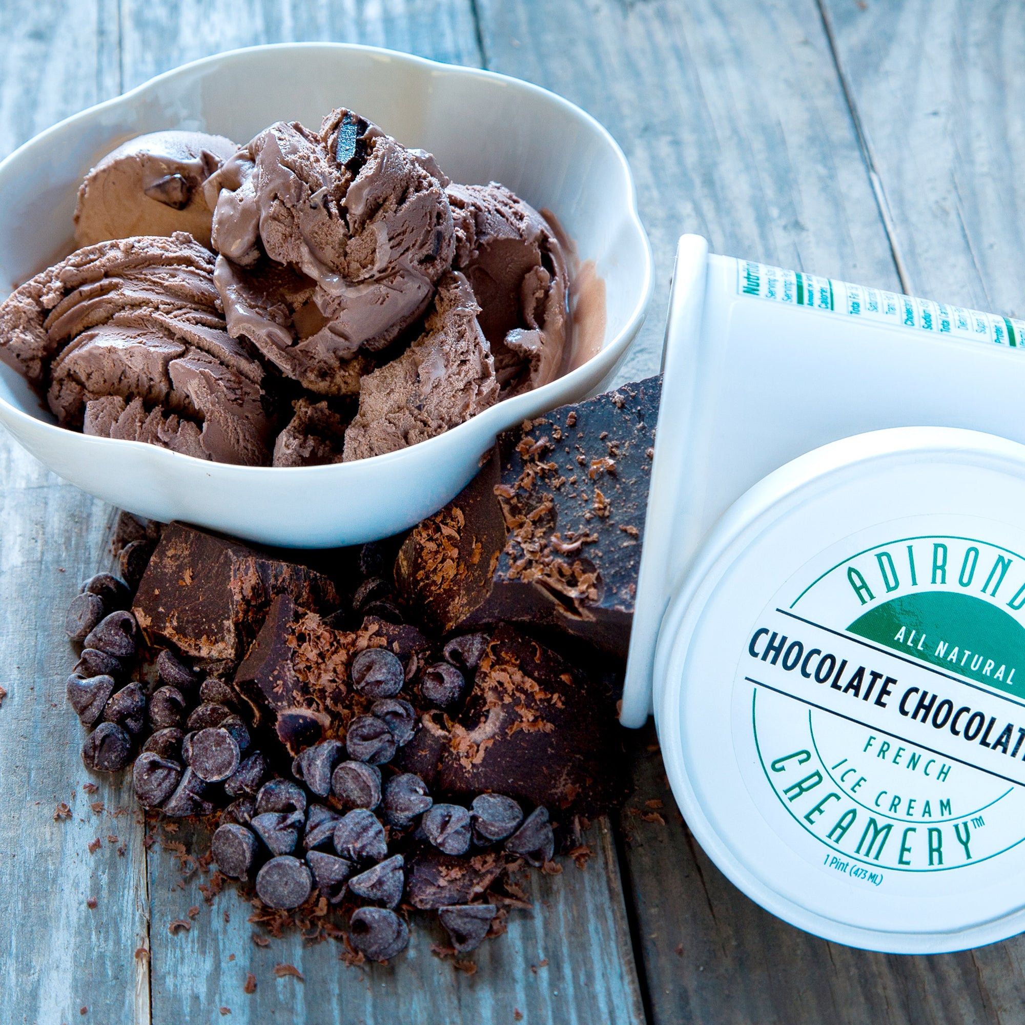 Bowl of chocolate chip ice cream with chocolate chips and a container of Adirondack Chocolate Creamery ice cream on a wooden surface.