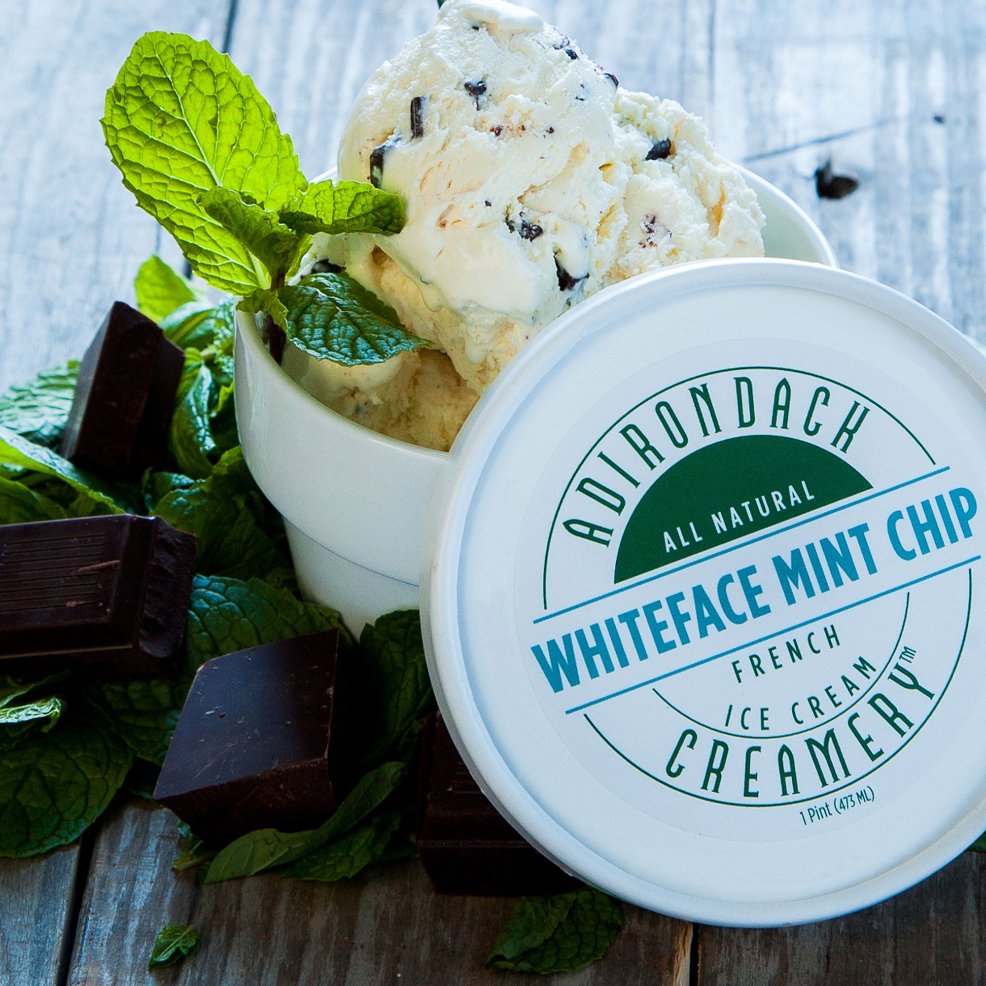 Adirondack All Natural Whiteface Mint Chip ice cream in a container with mint leaves and chocolate pieces on a wooden surface.