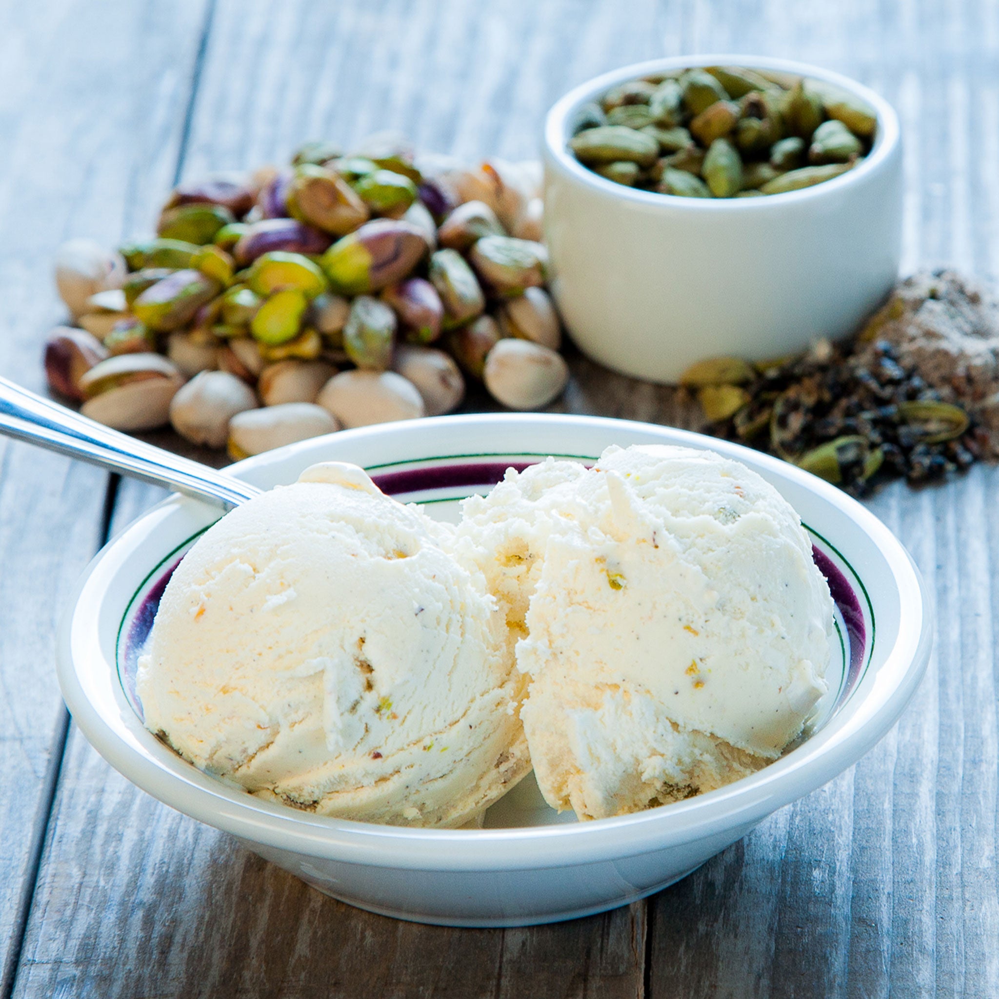 Two scoops of pistaccio ice cream in a white bowl with a spoon, surrounded by pistachios and a small container of pistachio nuts on a wooden surface.